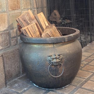 May include: A large, round, hammered brass pot with a dark patina finish, featuring a prominent lion head relief ornament holding a metal ring handle on the front, filled with several pieces of rough-cut, light brown firewood logs.