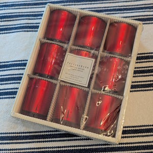 May include: A set of nine red mercury glass votive candle holders arranged in a square cardboard box. A central label reads Pottery Barn, mercury votives, set of nine, 1.9 oz, burn time 20 hours each. The votives have a reflective, metallic finish.