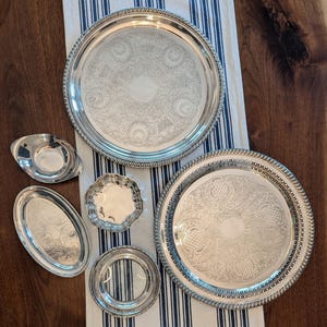 May include: A collection of six silver-toned serving pieces including two large round trays with intricate engraved patterns and rope-style edges, an oval dish, a small scalloped bowl, a plain oval tray, and a small round dish with a pierced rim.