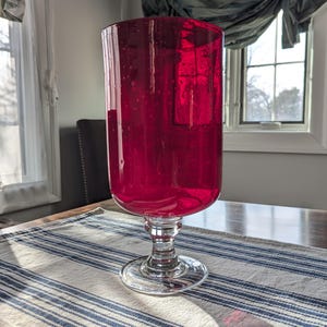 May include: A tall, ruby red glass vase with a wide, cylindrical body and a clear, stemmed base. The vase sits on a striped blue and white tablecloth. The glass has a slightly bubbly texture.