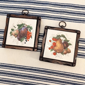 May include: Two square ceramic wall plaques set in dark brown wooden frames with metal hanging loops. Each plaque features a white center with a detailed botanical illustration of assorted fruits, including plums, red currants, pears, and raspberries.