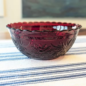 May include: A ruby red glass bowl with a scalloped rim and floral pattern. The bowl is made of pressed glass and sits on a blue and white striped tablecloth. The bowl's design features detailed flower and leaf motifs.