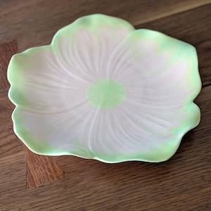 May include: A ceramic, flower-shaped dish with a pale pink center and a light green rim. The dish has a textured surface and a light green center. The dish is sitting on a wooden surface.