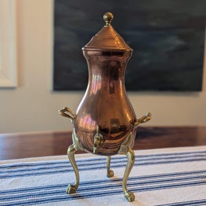 May include: A copper coffee pot with a lid and a brass spigot. The pot has a bulbous body and is supported by three brass legs. The pot has two brass handles and a brass knob on the lid.
