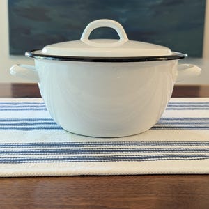 May include: A white enamel pot with a matching lid and two side handles. The pot features a smooth, glossy white finish with a thin, contrasting black rim around the top edge of the base and the lid. The lid has a central arched handle.