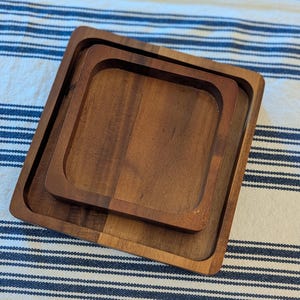 May include: Two square wooden trays with rounded interior corners, nested one inside the other. The trays feature a natural brown wood grain pattern with a smooth finish and raised edges, showcasing a simple, functional design for holding small items.