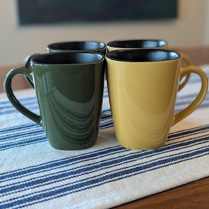 May include: Four ceramic mugs in shades of green and yellow, each with a black interior. The mugs have a square shape and curved handles. They are arranged on a white and blue striped tablecloth.