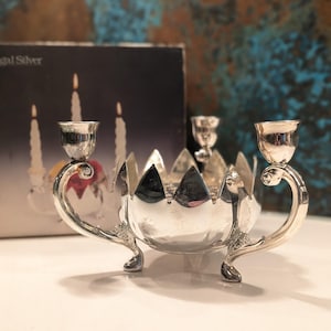 May include: A silver metal candle holder with two curved arms and a central bowl. The candle holder has a decorative design with a scalloped edge and three small feet. The background includes a box with images of candles.