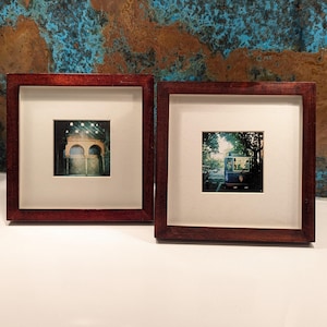 May include: Two framed square art prints. One print features an arched doorway with a decorative gate. The other print shows a street scene with a trolley car. Both prints are set in dark wood frames with white matting.