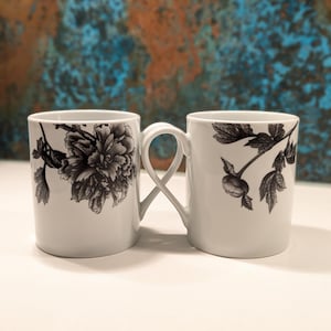 May include: Two white ceramic mugs with black floral designs. One mug features a large peony, the other a botanical illustration. The handles of the mugs are intertwined, creating a unique design. Ideal for coffee or tea.