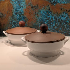 May include: Two white ceramic serving bowls with brown lids and wooden knobs. The bowls have small handles on either side. The background features a textured wall with blue and brown tones. The bowls are sitting on a white surface.