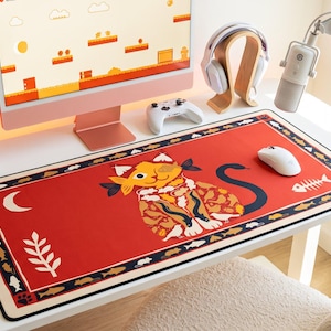 May include: A rectangular desk mat featuring a vibrant red background with a central illustration of a stylized cat wearing a patterned garment. The border is decorated with small fish, bones, and paw prints, with a white crescent moon and plant icon.