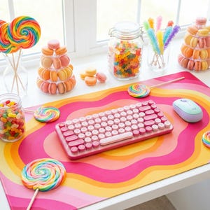 以下が含まれることがあります： ピンク色のワイヤレスキーボードとマウスが、虹色のデザインのカラフルなデスクマットの上に置かれています。お菓子、ロリポップ、マカロンが入った瓶が机を飾り、活気があり遊び心のあるワークスペースを作り出しています。