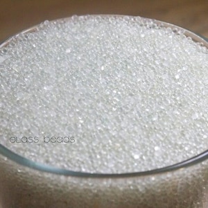 May include: A clear glass filled with small, transparent glass beads. The beads fill the glass to the top. The words "glass beads" are written in a light gray font on the side of the glass.