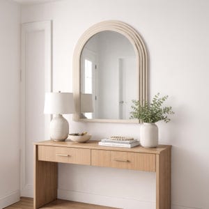Puede incluir: Una consola de madera clara con dos cajones, tiradores dorados y una lámpara blanca con una pantalla beige. Un espejo decorativo con la parte superior arqueada cuelga sobre la mesa. Un jarrón blanco con vegetación y objetos decorativos completan la escena.