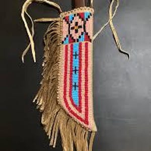 Könnte beinhalten: Eine handgefertigte Messerscheide mit einem Perlendesign in Rot, Blau und Beige. Die Scheide hat Fransen am Boden und Lederbänder. Der Messergriff ist oben sichtbar. Die Scheide ist etwa 25 cm lang.