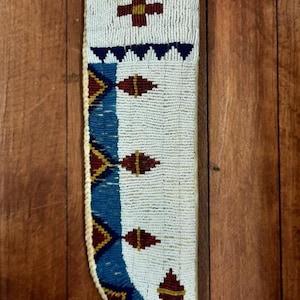 Könnte beinhalten: Eine mit Perlen besetzte Messerscheide mit weißem Hintergrund und blauen und kastanienbraunen geometrischen Mustern. Die Scheide hat ein Kreuz und Rautenformen. Die Scheide besteht aus kleinen Perlen und hat einen Trageriemen.