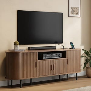 May include: A modern living room setup featuring a large black television mounted above a wooden entertainment center. The center has a soundbar, a stereo receiver, and a smartphone on a charging stand. Decorative plants and framed art complete the scene.
