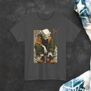 May include: Dark grey t-shirt with a detailed illustration of a figure with a cube-shaped head, wearing an orange jacket and green trousers. The artwork includes a lantern and decorative elements, creating a unique artistic design.