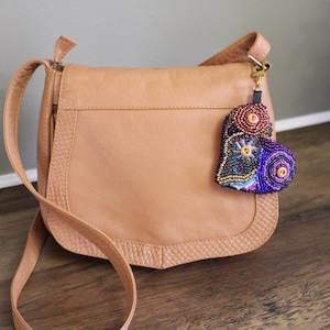 Puede incluir: Un bolso bandolera de cuero color canela con una correa larga. Un charm de cuentas con un cierre dorado cuelga del bolso. El charm presenta cuentas coloridas en tonos rojos, azules, morados y dorados.