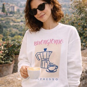 Puede incluir: Una sudadera blanca de manga larga con un estampado gráfico de una cafetera italiana azul y una taza de café sobre un fondo cuadrado beige claro. El texto "BUONGIORNO" está impreso en rosa arriba y la palabra "ESPRESSO" en azul debajo.