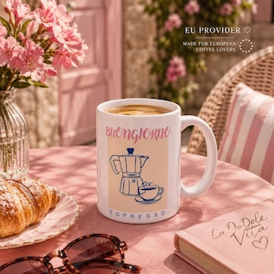 May include: A white ceramic mug featuring a graphic of a moka pot and a small cup of coffee. The text "Buongiorno" is printed in pink above the graphic, and the word "Espresso" is printed in blue below it. The mug has a curved handle.