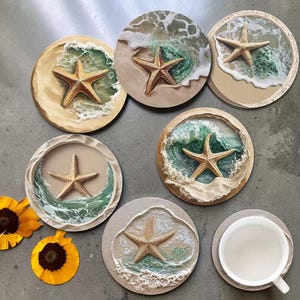Pode incluir: Seis porta-copos circulares com uma estrela-do-mar texturizada numa praia de areia com ondas a quebrar. Cada porta-copos apresenta um design de resina com tons de azul, verde e bege. Uma chávena de cerâmica branca está sobre um dos porta-copos.