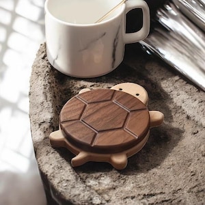 Puede incluir: Una taza de cerámica blanca con un asa negra y el texto "But First, Coffee" en negrita negra. La taza está sobre un fondo borroso. La taza mide aproximadamente 10 cm de altura.