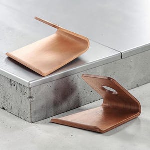 Op de afbeelding: Twee houten telefoonstandaards met een gebogen ontwerp. De standaards zijn gemaakt van lichtbruin hout en hebben een gladde afwerking. Eén standaard staat rechtop en de andere ligt plat. De standaards staan op een betonnen oppervlak.
