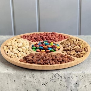 Könnte beinhalten: Ein runder Servierteller aus hellem Holz mit fünf Fächern. Der Teller ist mit verschiedenen Snacks gefüllt, darunter Erdnüsse, Cashewnüsse, Walnüsse, Mandeln und bunte Süßigkeiten. Ideal zum Servieren von Vorspeisen oder Snacks.