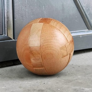 Può includere: Un puzzle a sfera in legno con un design segmentato. La palla è realizzata in legno di colore chiaro e ha una finitura liscia e lucida. I segmenti sono disposti per creare una forma sferica, con cuciture visibili. Il puzzle è progettato per essere smontato e rimontato.