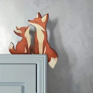 May include: Two wooden fox figurines, one large and one small, are perched on a light blue surface. The foxes are primarily orange with white accents, featuring closed eyes and a seated pose. The background is a textured gray wall.