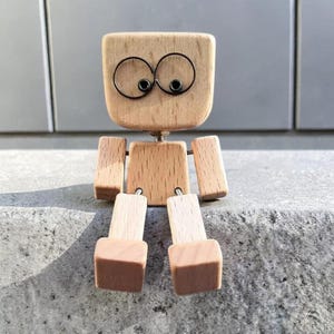Op de afbeelding: Een houten robotfiguur met een vierkante kop en rechthoekig lichaam en ledematen. De ogen zijn gemaakt van zwart draad. De voeten hebben een bruine afwerking. De robot zit op een grijs oppervlak.