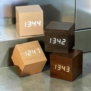 May include: Four cube-shaped digital clocks with wood-grain finishes in light, medium, and dark brown tones. Each clock features a glowing white LED display showing different times: 13:44, 13:42, 12:47, and 13:43, presented in a simple block design.