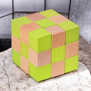 Può includere: Un puzzle a cubo in legno con un motivo a scacchiera. Il cubo è composto da blocchi più piccoli in due colori: verde chiaro e legno naturale. Il puzzle è appoggiato su una superficie di pietra.