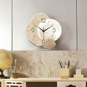 Puede incluir: Un reloj de pared moderno con un diseño único. La esfera del reloj es de color beige claro con números y manecillas negras. El reloj tiene forma circular con un diseño similar a la luna en el centro y una forma de piedra alrededor.