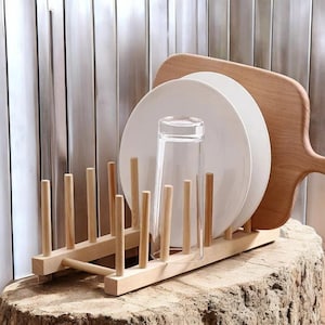 Puede incluir: Un escurreplatos de madera con clavijas verticales sostiene platos blancos y un vaso transparente. Una tabla de cortar de madera descansa detrás de los platos. El estante se encuentra sobre un tocón de árbol, con un fondo metálico.