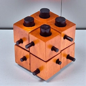 May include: A wooden puzzle cube with a natural wood finish. The cube is made up of smaller blocks, each with a dark brown peg and a cylindrical black peg. The puzzle is designed to be manipulated and solved.