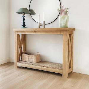 Puede incluir: Una consola de roble claro con un diseño en forma de X en los laterales. Una cesta tejida se encuentra en el estante inferior. La mesa está coronada con una lámpara verde, velas y un jarrón de flores. La mesa mide 122 cm de largo.