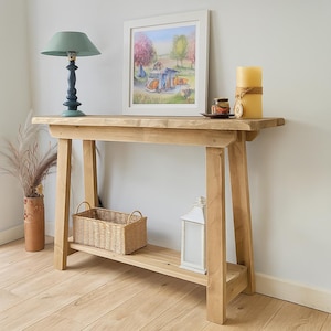 May include: A light-colored wooden console table with a lower shelf, holding a woven basket and a white lantern. A framed landscape painting, a candle, and a lamp with a teal shade are on the tabletop. The table is against a white wall.