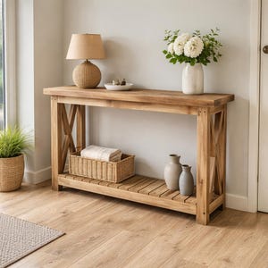 May include: A light brown wooden console table with an X-shaped design on the sides. The table has a lower shelf with a woven basket, two vases, and a lamp with a beige shade. A vase of white flowers sits on top.