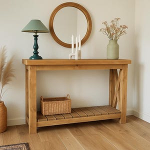 Può includere: Una consolle in legno chiaro con un ripiano inferiore e un cestino in vimini. Una lampada verde e uno specchio rotondo sono sopra. Un vaso con fiori secchi e un portacandele bianco sono sul tavolo.