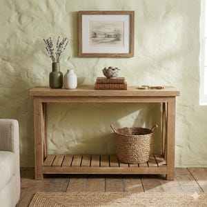 Peut inclure: Une console en bois clair avec une étagère inférieure et un panier en osier. La table est ornée d'une estampe encadrée, de livres, d'un bol décoratif et de vases avec des fleurs séchées. Le mur est vert clair.