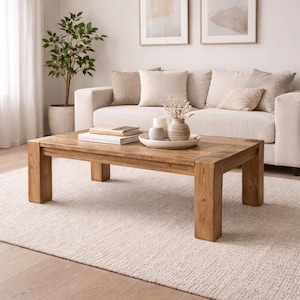 Puede incluir: Una mesa de centro rectangular de madera con acabado natural. La mesa tiene un diseño sencillo y robusto con patas gruesas y una superficie espaciosa. La mesa está en un salón con un sofá blanco, cojines y objetos decorativos.