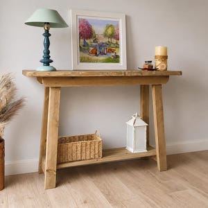 May include: A rustic wooden console table with a rectangular top and a lower shelf. The table features four angled legs and a natural wood grain finish. It is styled with a light blue lamp, a framed landscape print, a woven basket, and a white lantern.