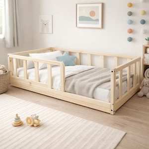 Op de afbeelding: Een licht houten peuterbed met veiligheidsrails. Het bed is opgemaakt met wit beddengoed, een grijze deken en een blauw stervormig kussen. De kamer is ingericht met een ingelijste foto en kinderspeelgoed.