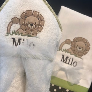 May include: White hooded towel with a green and white checkered border. The towel has an embroidered lion with the name "Milo" underneath. A matching white washcloth with a green and white checkered border and an embroidered lion with the name "Milo" underneath.