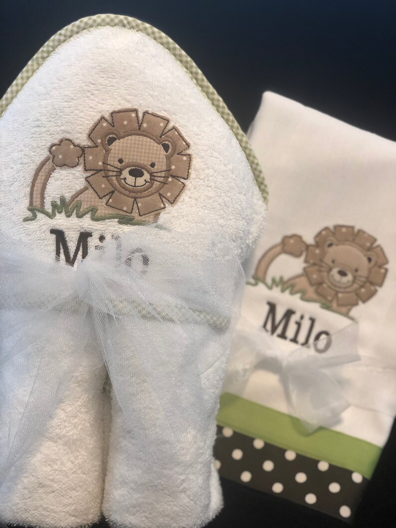 May include: White hooded towel with a green and white checkered border. The towel has an embroidered lion with the name "Milo" underneath. A matching white washcloth with a green and white checkered border and an embroidered lion with the name "Milo" underneath.
