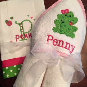 May include: A white hooded towel with a pink gingham trim and a green frog with a pink bow embroidered on it. The towel is tied with a white ribbon and the name "Penny" is embroidered on the towel.