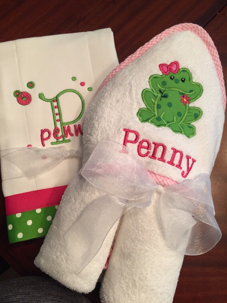 May include: A white hooded towel with a pink gingham trim and a green frog with a pink bow embroidered on it. The towel is tied with a white ribbon and the name "Penny" is embroidered on the towel.
